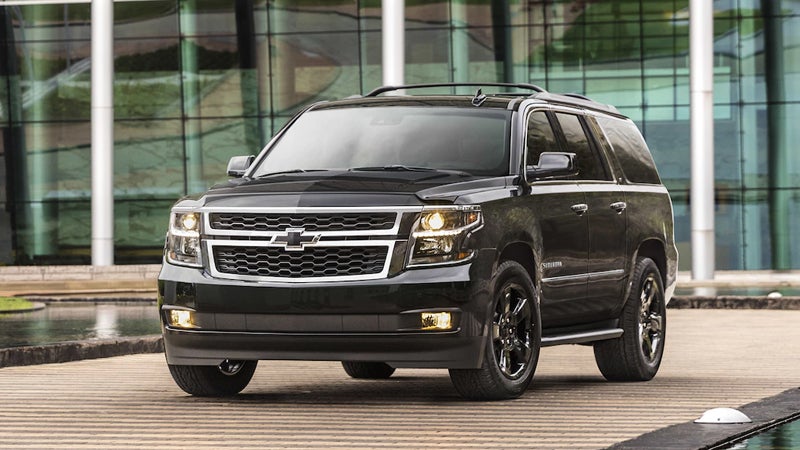 A blacked-out Chevrolet Suburban parked in front of a modern glass building, featuring black wheels and dark trim.