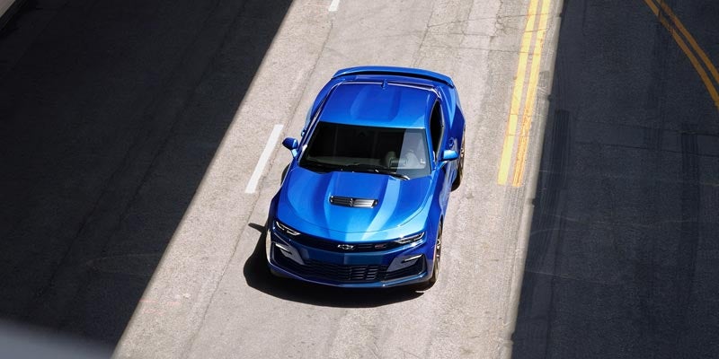 A striking blue 2021 Chevrolet Camaro seen from above, driving through a city street with sunlight casting sharp shadows.