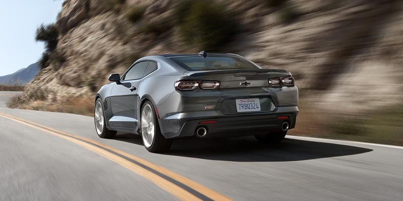 A silver Chevrolet Camaro driving through a mountain road, showing its aggressive rear styling and dual exhaust.
