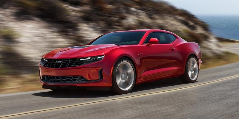 A red Chevrolet Camaro driving fast along a coastal highway, emphasizing its sculpted body and muscular stance.