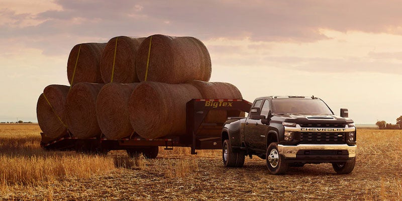 Chevrolet Silverado HD towing a large trailer loaded with hay bales in an open field at sunset.