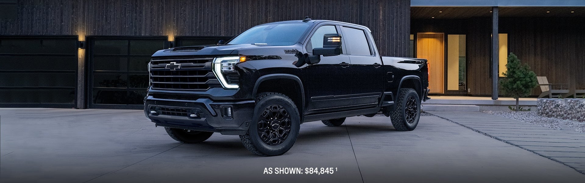 Front-side view of a black 2024 Chevrolet Silverado HD parked in front of a modern home at dusk