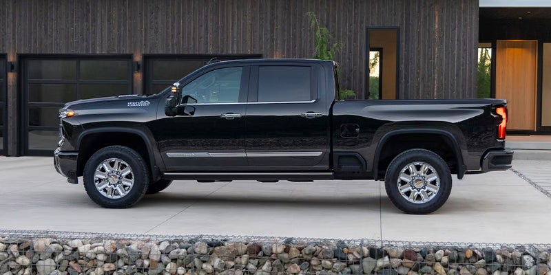 Side profile of the 2024 Chevrolet Silverado HD parked in front of a modern house, showcasing its extended cab and polished wheels.